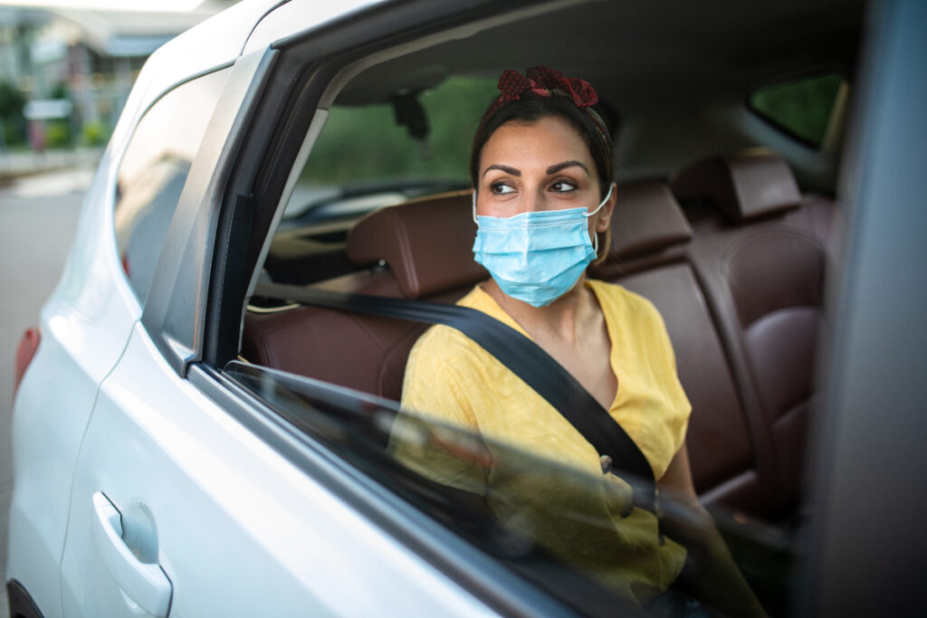 A woman wearing a yellow shirt and face mask rides in the backseat of an Uber car while looking wistfully out the window.